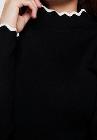 Svart ribbat polokrage-tröja med vit scallopkant vid halsringningen och ärmarna, som framhäver dess texturerade tyg och figursydda design.
