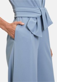 Világoskék overall, amely illeszkedő felsőrészből, széles szárból, masnival díszített övből és praktikus zsebekből áll. Simaság jellemzi a szövet textúráját.