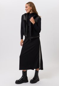 Black sleeveless vest with textured shoulders over a black long-sleeve dress, paired with chunky black ankle boots. Smooth and fitted materials.