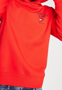 Rode hoodie van zacht materiaal met een logo aan de voorkant in zwart en meerkleurige accenten. Het heeft een ontspannen pasvorm en ribgebreide manchetten.