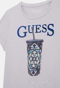 Světle fialové tričko má tmavě modrý nápis "GUESS", grafiku s motivem vzorovaného hrnku na nápoj a vyšitý detail "Los Angeles" blízko spodního lemu.
