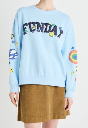 Lichtblauwe trui met kleurrijke "FUNDAY" tekst, glimlachende gezichten, bloemen, wolken en regenboogontwerpen, gedragen met een bruine ribfluwelen rok.