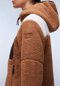 Bruine fleecejas met witte en bruine accenten, een gestructureerd oppervlak, capuchon en een Noors vlagpatch op de mouw. Inclusief een ritszak.