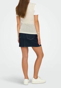 Knit beige vest with a perforated design, worn over a white t-shirt. Paired with a dark denim skirt and white sneakers, displaying a casual style.