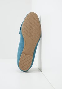 Chaussure plate en suede bleu avec un bout arrondi et une semelle en caoutchouc texturée. Dotée d'une sangle noire sur le cou-de-pied et d'un design léger.