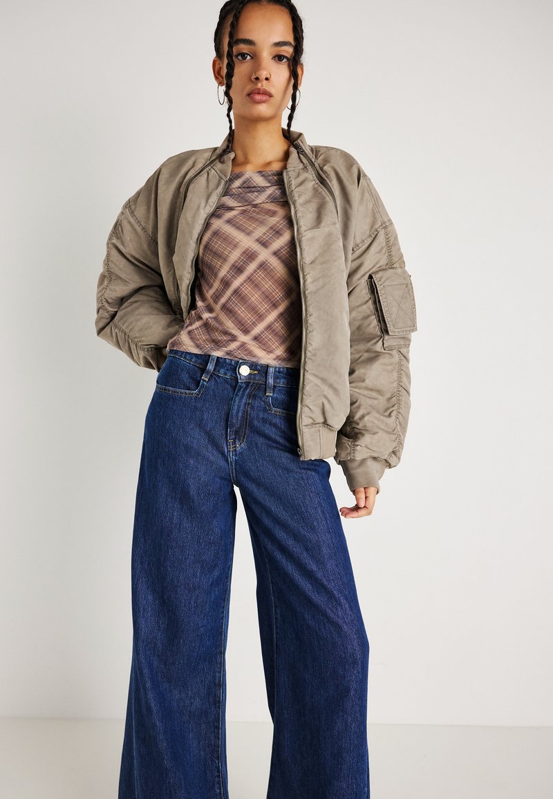 Casaco de bombardeiro caqui com textura acolchoada, combinado com uma blusa de xadrez bege e calças de ganga azul de perna larga. Design simples e contemporâneo.