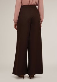 Breda byxor i brun väv med hög midja. Slät textur med en strukturerad passform. Bakifrån visar skräddarsydda sömmar och rena linjer.