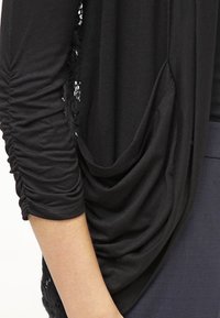 Black draped cardigan featuring ruched long sleeves, a soft texture, and a lace accent along the edges. Designed for a relaxed fit.