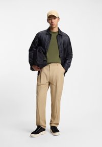 Sort letvægtsjakke, grøn sweater, beige bukser og sorte sneakers med hvide såler. Beige kasket med logo. Teksturen varierer på tværs af stoffer.