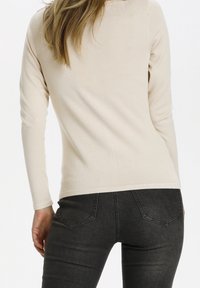 Beige långärmad topp med figursydd design och slät textur, kombinerad med mörkgrå jeans med bakfickor och diskret sömdekoration.