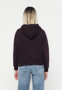 Haut court en tissu violet foncé avec une capuche à cordon. Présente des manches longues et des poignets côtelés, associé à un jean en denim bleu clair.