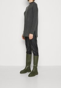 Grå ribbad tröja med sidofickor, kombinerad med glansiga svarta konstläderbyxor och knähöga olivgröna stövlar. Släta texturer och figurnära design.