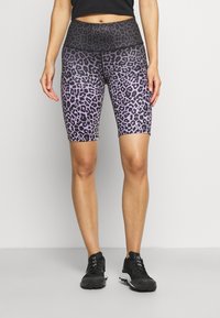 Kobieta w purpurowych i czarnych szortach rowerowych w panterkę z wysokim stanem, czarnym topie na brzuch i czarnych butach sportowych stoi na szarym podłodze.