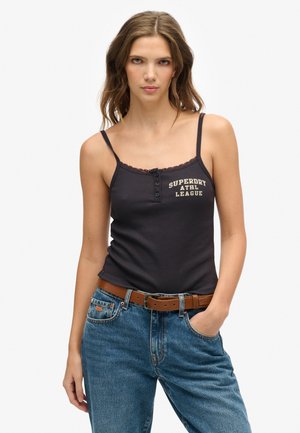Femme portant un débardeur noir orné de dentelle avec le texte "SUPERDRY ATHL LEAGUE", un jean bleu et une ceinture marron, posant avec une main dans la poche.