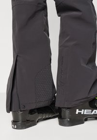 Fekete vízálló sínadrág texturált alsó panellel, cipzárral a szegélyen és márkajelzéssel az oldalon, fekete sícipőkkel párosítva.