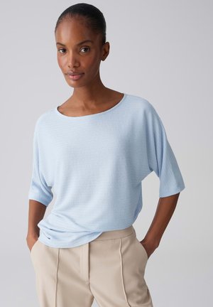 Frau mit zurückgebundenem Haar, die ein hellblaues, strukturiertes Kurzarmoberteil und beige Hosen trägt, stehend mit den Händen in den Taschen vor grauem Hintergrund.