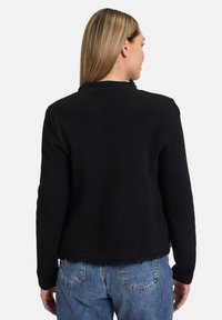 Černá mikina se třásněmi na spodním lemu, s dlouhými rukávy a kulatým výstřihem, nošená přes modré džíny.