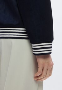 Veste côtelée marine avec poignets rayés blancs et marines, présentant une coupe décontractée. Une main apparaît au niveau du poignet, montrant le ton et la texture de la peau.