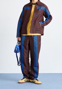 Completo sportivo marrone a fantasia con strisce blu, abbinato a una maglietta gialla e sneaker gialle. Una piccola borsa blu è tenuta in una mano.