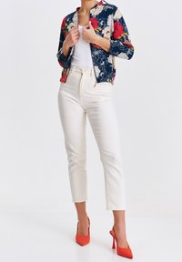 Chaqueta bomber floral en azul marino, con flores rojas y crema. Combina con pantalones blancos hasta el tobillo y tacones rojos puntiagudos. Tela de textura suave.