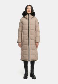 Beige Steppjacke mit langem, geradem Schnitt, gestepptem Textil, schwarzem Kunstfellkragen, vorderem Reißverschluss und Seitentaschen. Getragen mit schwarzen Hosen und Stiefeln.