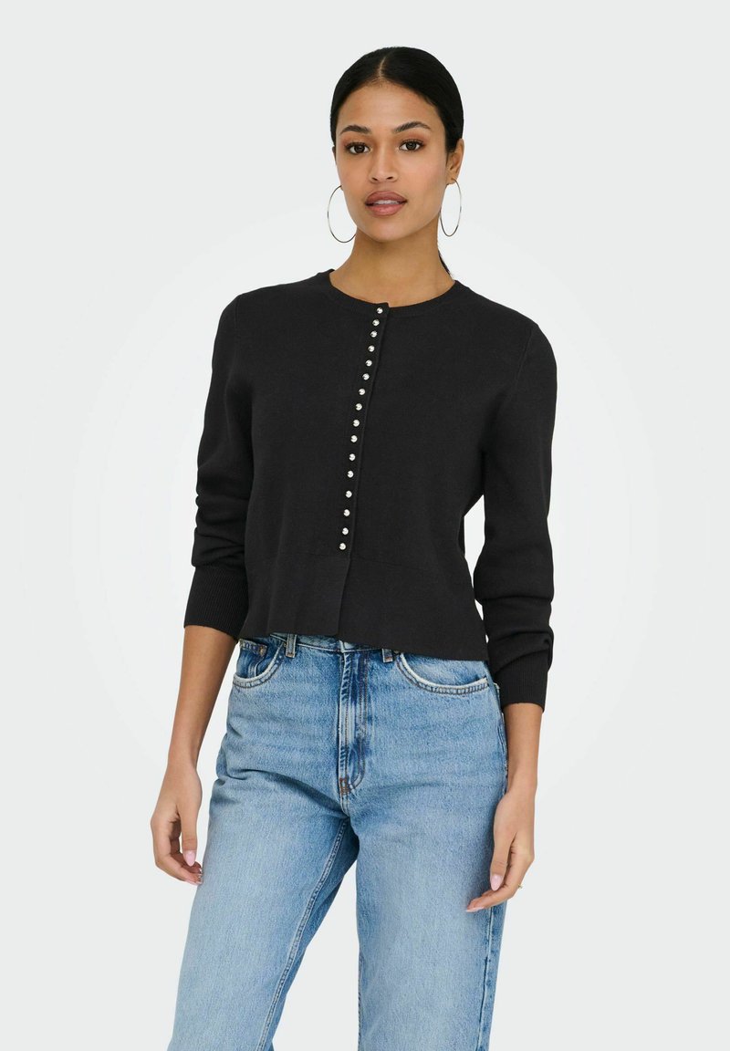 Femme portant un cardigan noir boutonné et un jean taille haute bleu clair, debout devant un fond clair uni.