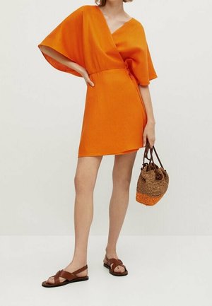 Femme portant une robe portefeuille orange à manches larges, tenant un sac tissé bicolore et portant des sandales plates marron.