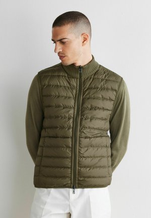 Man draagt een olijfgroene doorgestikte bodywarmer met rits over een shirt met lange mouwen, staat tegen een witte muur en kijkt naar beneden opzij.