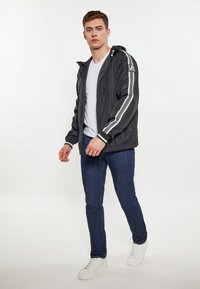 Giacca leggera nera con cappuccio, caratterizzata da strisce bianche lungo le maniche. Indossata sopra una maglietta bianca, abbinata a jeans scuri e sneaker bianche.