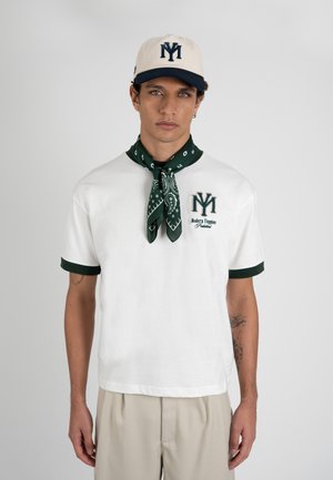 Ung mand iført hvid T-shirt med grøn kant, mørkegrønt mønstret halstørklæde, beige bukser og hvid kasket med mørkeblå logo.