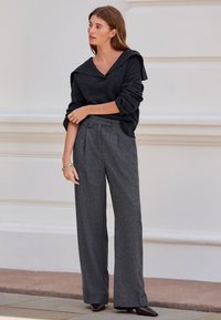 Schwarzer Strickpullover mit weiter Kapuze und umgeschlagenen Ärmeln, kombiniert mit hoch taillierter, grauer Hose mit Muster und zwei Knöpfen.