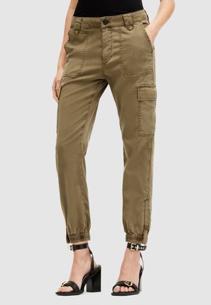 Kobieta ubrana w oliwkowe spodnie typu cargo z kieszeniami naszywanymi z przodu i elastycznymi mankietami, w połączeniu z czarnymi sandałami na wysokim obcasie.