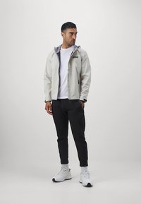 Világosszürke kapucnis dzseki fehér inggel, sötét nadrággal párosítva. A dzsekien logó és cipzáras zsebek találhatók, semleges háttér előtt.