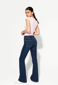Rosa enganzt geschnittenes T-Shirt, kombiniert mit dunkelblauen Schlagjeans. Die Jeans haben Gesäßtaschen und kontrastfarbene Nähte. Die Frau steht im Profil.