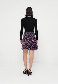 Femme debout de dos, portant un haut noir à manches longues, une jupe fleurie violette et des sandales noires à talons hauts sur fond blanc.