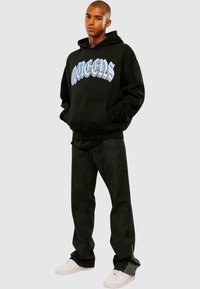 Sort hættetrøje med "QUEENS" i lyseblå skrift, parret med mørke denimbukser og hvide sneakers, minimalistisk design.
