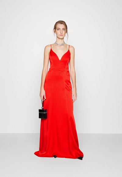 Donna che indossa un abito senza maniche in satin rosso con una profonda scollatura a V, una collana d'argento, mentre tiene una piccola borsa nera su uno sfondo semplice.