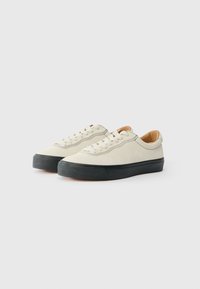 Gai beigi zamšādas tenisi ar melniem gumijas zole, apaļu purngalu un tonālā šuvuma detaļām. Iespējas ar saites dizainu un minimālistisku stilu.