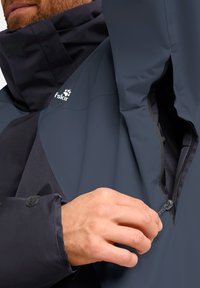 Giacca impermeabile navy con colletto alto, tasca con zip e polsini in velcro. Presenta una superficie liscia e elementi di design ergonomico.