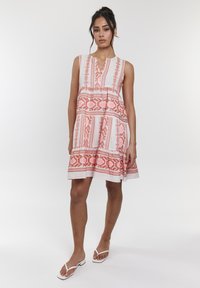 Ärmelloses Kleid aus einem leichten Stoff mit rosa und korallenfarbenen geometrischen Mustern, das über einen V-Ausschnitt und einen schwingenden Saum verfügt. Getragen mit flachen weißen Sandalen.