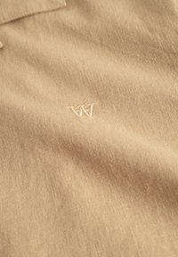 Polo beige en tissu texturé avec une finition douce. Il présente un logo "W" brodé sur la poitrine, mettant en avant des détails subtils.
