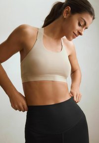 Beige sportbh med breda band och en band prytt med logo. Slät material med en figurnära passform, kombinerad med svarta högmidjade leggings.