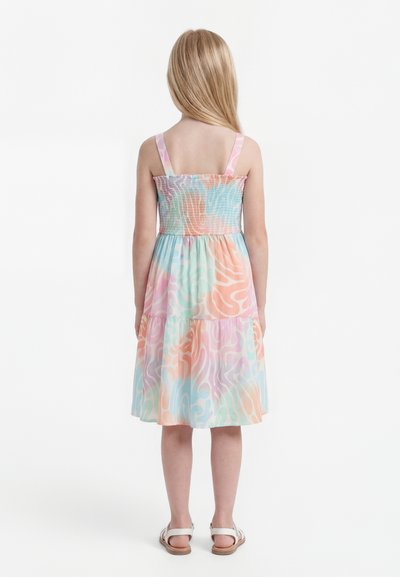 Κορίτσι με μακριά ξανθά μαλλιά, ντυμένο με ένα αμάνικο, παστέλ, στρωματοποιημένο φόρεμα και λευκά σανδάλια, που στέκεται γυρισμένο με την πλάτη.