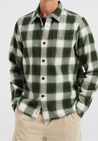 Green and white plaid flannel shirt with dark buttons, a single chest pocket, and a textured fabric. Paired with light-colored corduroy pants.