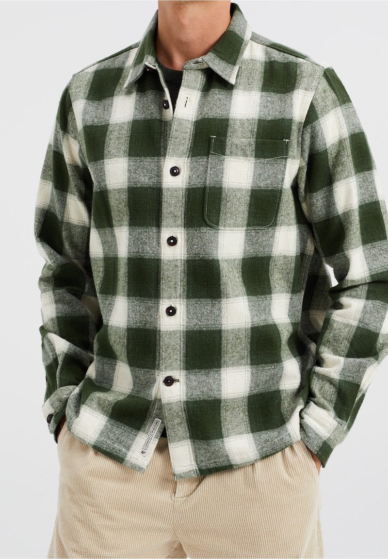 Green and white plaid flannel shirt with dark buttons, a single chest pocket, and a textured fabric. Paired with light-colored corduroy pants.