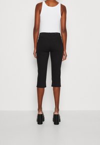 Osoba v černých zkrácených kalhotech, bílém bezsleeve topu a černých platformových botách, stojící zády k jednoduchému pozadí.