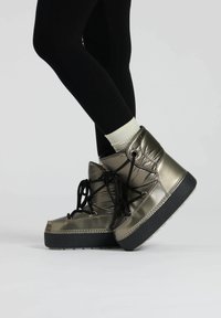 Stivaletti metallici verde oliva con design imbottito, lacci neri e suole in gomma nera testurizzata, indossati con leggings neri e calzini bianchi.