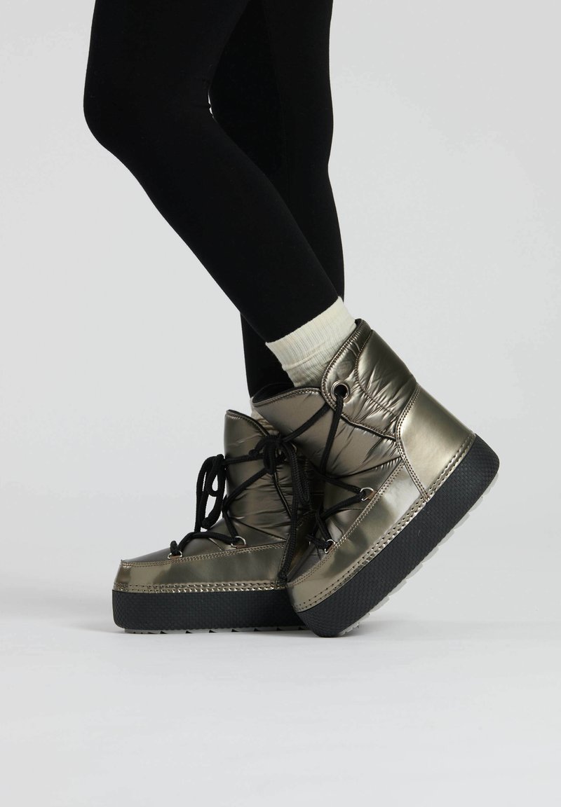 Stivaletti metallici verde oliva con design imbottito, lacci neri e suole in gomma nera testurizzata, indossati con leggings neri e calzini bianchi.