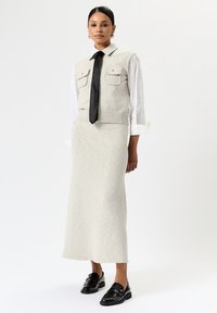 Gilet tricot grigio chiaro con due tasche sul petto, indossato sopra una camicia bianca e una gonna grigia abbinata. Una cravatta nera e mocassini lucidi completano il look.