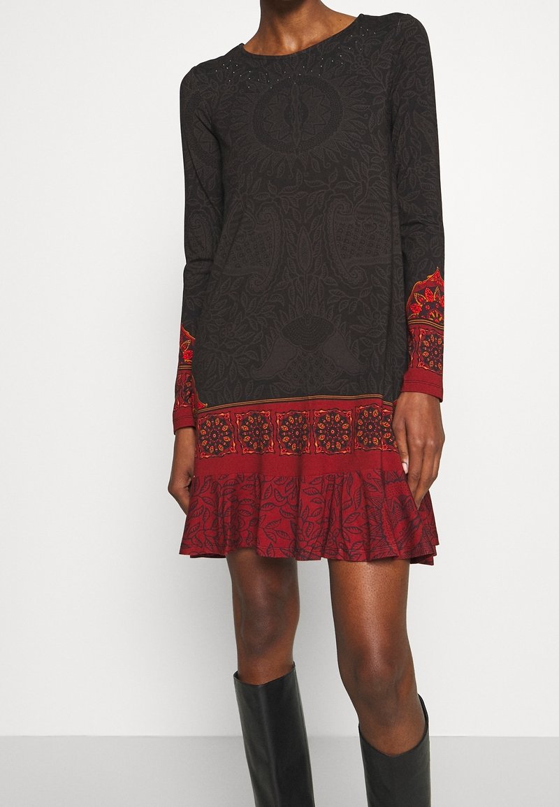 Femme portant une robe noire et rouge à manches longues avec des motifs floraux et cachemire, et des bottes noires montantes jusqu'aux genoux.